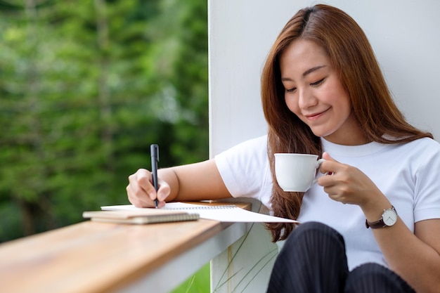 Een mooie jonge Aziatische vrouw die koffie drinkt en in de buitenlucht op een notitieboekje schrijft