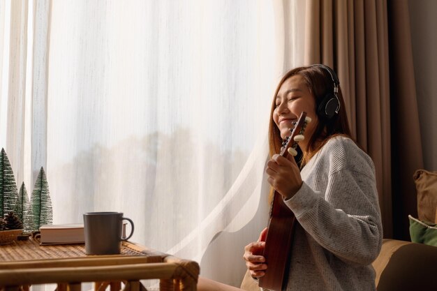 Een mooie jonge Aziatische vrouw met koptelefoon speelt graag thuis een ukelele