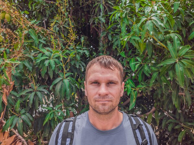 Een mooie jonge blanke man in een grijs shirt met een rugzak in de buurt van groene mangoboom Reisconcept met kopieerruimte