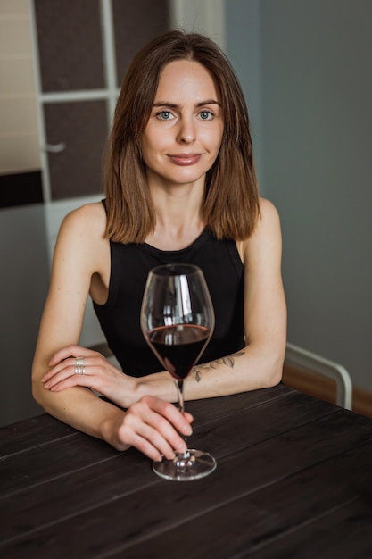 Een mooie jonge blanke vrouw zit aan de houten tafel in de keuken met een glas rode wijn in haar handen