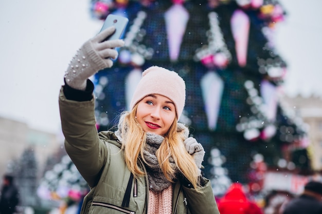 Een mooie jonge blonde vrouw lachend naar een telefoon voor een foto op de kerstmarkt