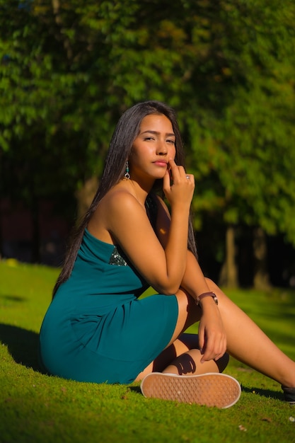 Een mooie jonge brunette Latina met lang steil haar, gekleed in een strakke groene jurk. Portret van een meisje, zittend op het gras in een park met bomen in de zomer
