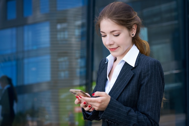 Een mooie jonge brunette meisje in een shirt en jas staat tegen de achtergrond van een kantoorgebouw met een telefoon in haar handen op een zonnige dag