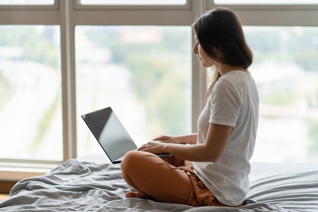 Een mooie jonge brunette meisje werkt op de laptop zittend op een bed bij een panoramisch raam. Winkelen op internet.