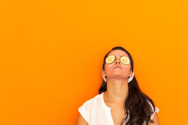 Een mooie jonge Latijnse vrouw met een schoonheidsmasker op haar gezicht en stukjes komkommer in haar ogen op een oranje achtergrond. Zelfzorgconcept