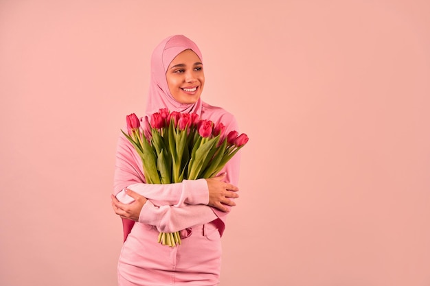 Een mooie jonge stijlvolle moslimvrouw in een roze hijab en kleding houdt een boeket tulpen vast en glimlacht terwijl ze naar de zijkant kijkt