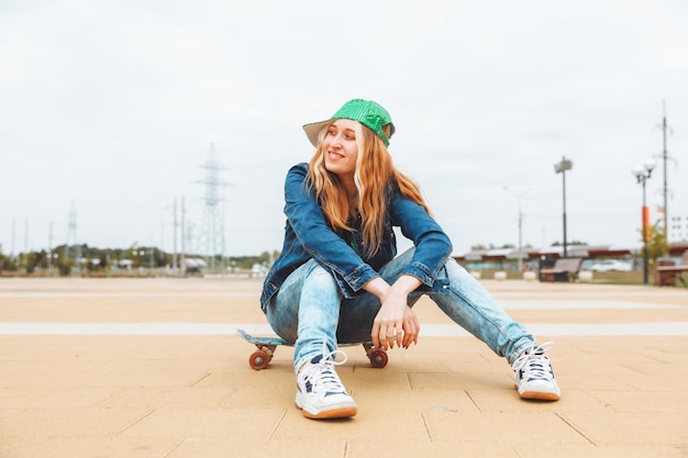 Een mooie jonge tienermeisje in een pet en denimjas zit over een skateboard generatie z