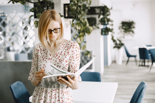 Een mooie jonge vrouw die binnen in een café staat en wat documenten leest. Vrouw met bril en bloemenjurk