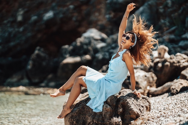 Een mooie jonge vrouw gekleed in een blauwe jurk heeft plezier op het strand op de zonnige dag.