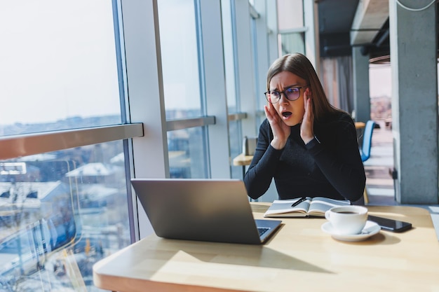 Een mooie jonge vrouw in een beige jas en bril las het nieuws op een laptop in internetbronnen en was geschokt door wat ze las