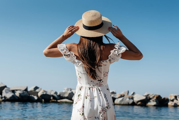 Een mooie jonge vrouw in een hoed en een lichte jurk met haar rug loopt langs de oceaankust tegen de achtergrond van enorme rotsen op een zonnige dag Toerisme en vakantiereizen