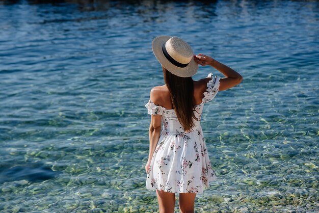 Een mooie jonge vrouw in een hoed en een lichte jurk met haar rug loopt op een zonnige dag langs de oceaankust tegen de achtergrond van enorme rotsen. Toerisme en vakantie reizen.