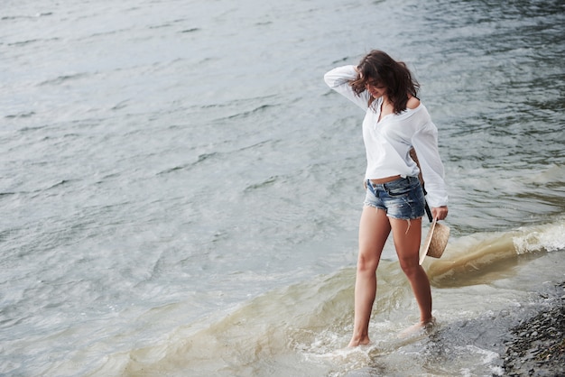 Een mooie jonge vrouw in een hoed en met een rugzak loopt speels door het water. Een warme zomerdag is een geweldige tijd voor avontuur en avontuur in de natuur