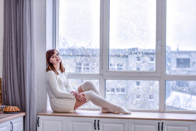 Een mooie jonge vrouw in een witte gebreide truijurk en lange legging zit op een brede vensterbank bij het raam. op zoek naar de afstand, verdrietig, eenzaamheid concept. kopieer ruimte. witte interieurs