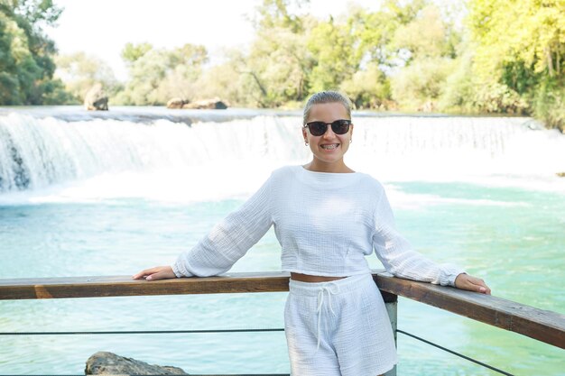 Een mooie jonge vrouw in een witte korte broek en een shirt tegen de achtergrond van een bergwaterval tegen de achtergrond van een dicht bos Prachtig landschap