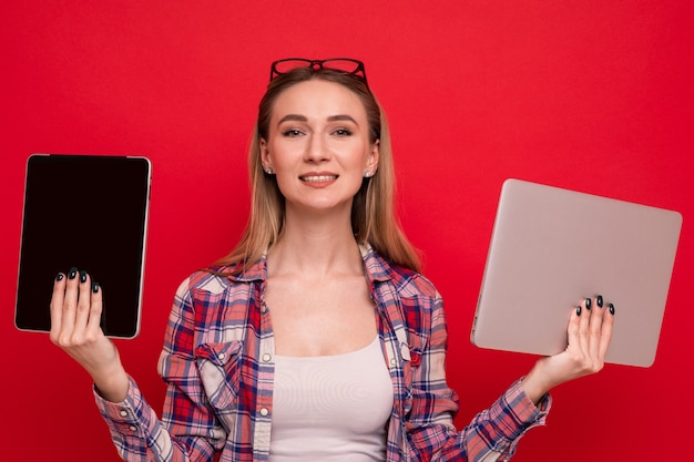 Een mooie jonge vrouw in stijlvolle kleding heeft een tablet en een laptop op een rode achtergrond