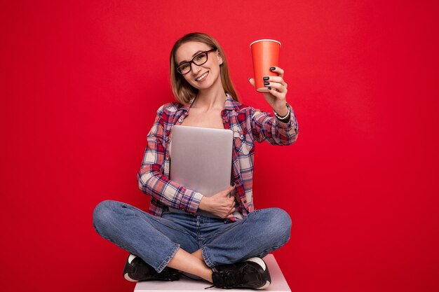 Een mooie jonge vrouw in stijlvolle kleding zit met een kartonnen beker en een laptop op een rode achtergrond