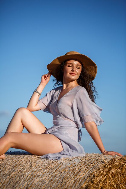 Een mooie jonge vrouw met een hoed en een zomerjurk zit op een hooischoof in een veld. Landelijke natuur, tarweveld