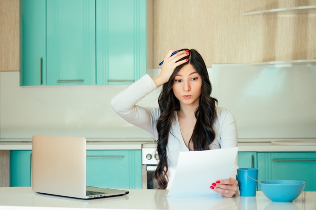 Een mooie jonge vrouwelijke student (freelancer) in een witte jurk en zwart haar werkt thuis met een laptop en papieren in een turquoise keuken. Het idee om het probleem en de deadline op te lossen