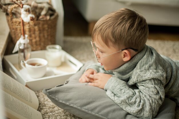 Een mooie kleine jongen in een gebreide trui leest bij het raam met een mok hete thee. knus. herfst. valstemming.