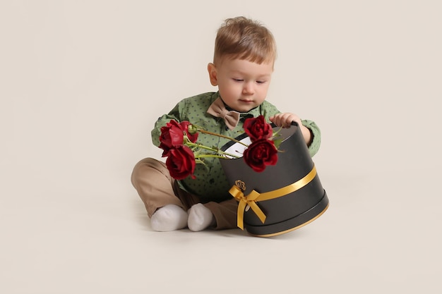 een mooie kleine jongen in een groen shirt zit op een witte achtergrond naast een doos met rode rozen