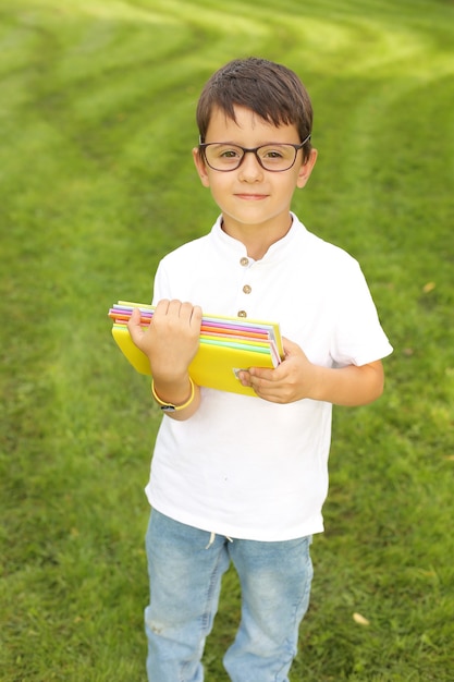 een mooie kleine jongen staat in het park in een wit T-shirt met een bril met notitieboekjes