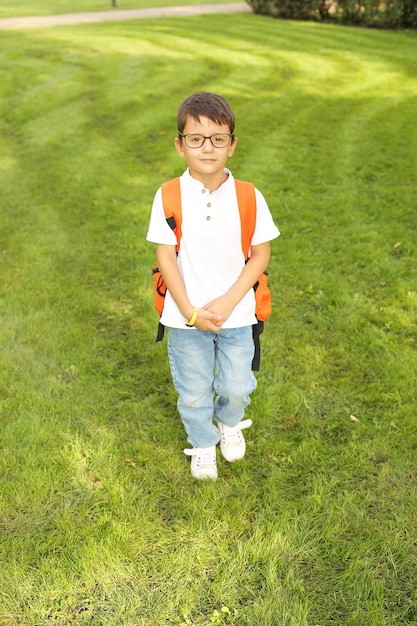 een mooie kleine jongen staat in het park in een witte T-shirtjeans met een oranje rugzak