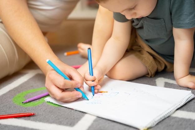 Een mooie kleine peuterjongen van twee of drie jaar oud met papa tekent met stiften in het album in de kinderkamer thuis, zittend op de vloer Tijd doorbrengen met kinderen Selectieve aandacht