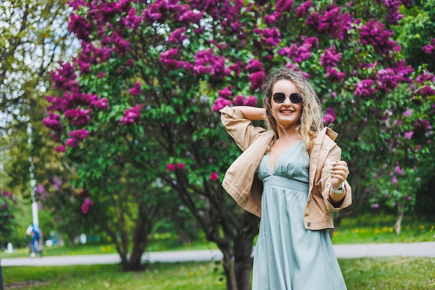 Een mooie krullende vrouw in een zomerjurk rent en verheugt zich dat ze een zonnebril draagt tegen de achtergrond van een paarse lila struik