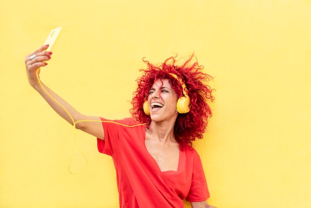 Een mooie lachende latijnse vrouw met rood afrohaar luistert naar muziek met gele koptelefoon en neemt een selfie met een gele smartphone op een gele achtergrond