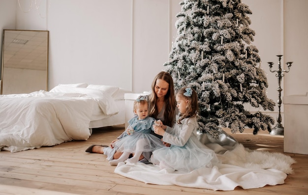 Een mooie moeder en haar dochters in feestelijke blauwe jurken zitten bij de kerstboom en spelen