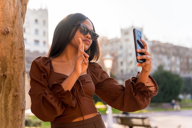 Een mooie natuurlijke jonge Afrikaanse vrouw in een park met zonnebril kijkend naar de telefoon bij zonsondergang