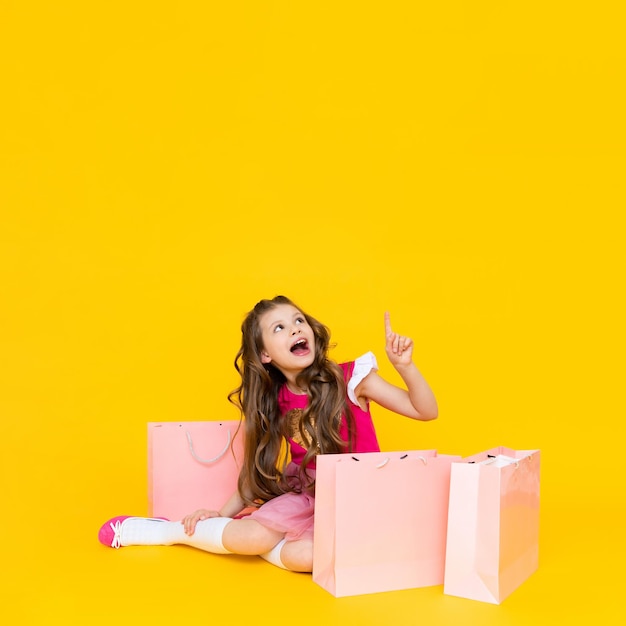 Een mooie prinses met winkelen zit op een gele geïsoleerde achtergrond Het kleine meisje wijst omhoog naar uw advertentie en glimlacht breed Verkoop en winkelen voor kinderen Ruimte kopiëren