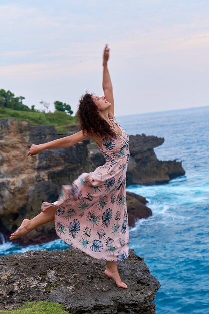 Een mooie slanke vrouw in een lange jurk poseert op een rots op een klif boven de oceaan