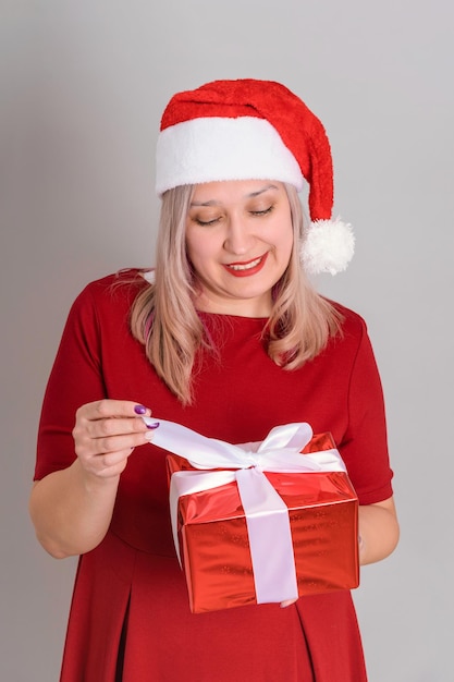 Een mooie volwassen vrouw met een kerstmuts en een rode jurk opent een geschenkdoos en lacht liefjes