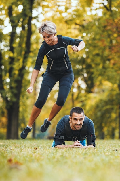 Een mooie vrolijke vrouw en haar partner zijn aan het sporten in het park, gekleed in een zwart pak met een EMS elektronische simulator om hun spieren te stimuleren.