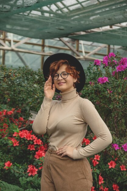 Een mooie vrouw in brillen en hoed die tussen de groene planten en bloemen glimlacht.