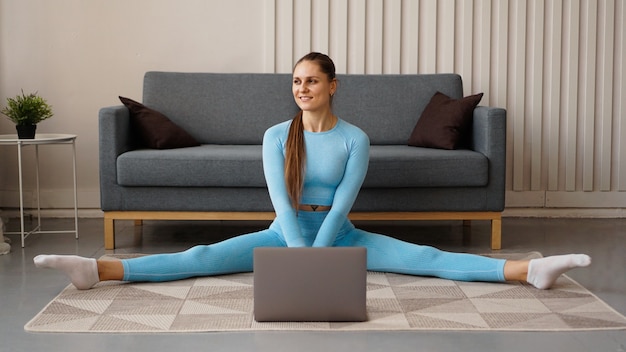 Een mooie vrouw in een blauw trainingspak strekt zich thuis uit voor een laptop. Ze zit in een dwarse koord.