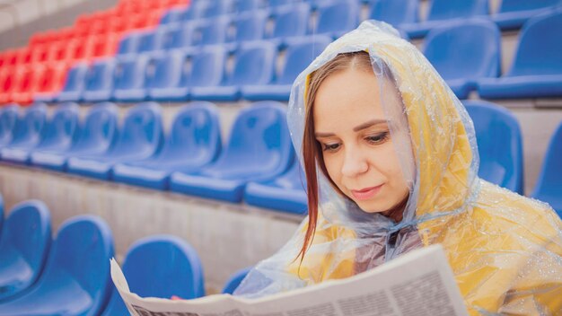 Een mooie vrouw in een hoodie en een plastic regenjas leert het laatste nieuws door tijdens de regen een krant te lezen die op het podium in een leeg stadion zit