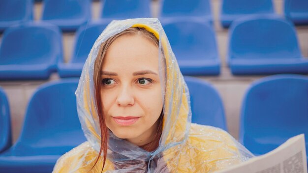 Een mooie vrouw in een hoodie en een plastic regenjas leert het laatste nieuws door tijdens de regen een krant te lezen die op het podium in een leeg stadion zit