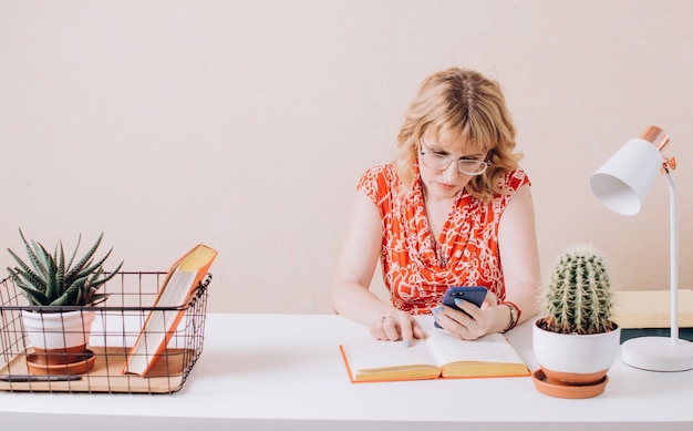 Een mooie vrouw in een rode blouse zit aan tafel en kijkt aandachtig naar de telefoon en het boek