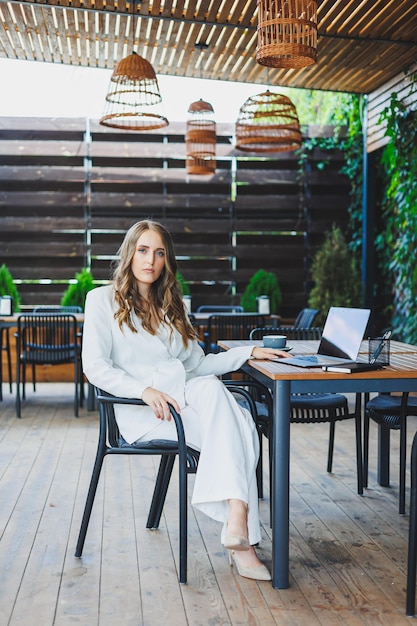 Een mooie vrouw in een wit klassiek pak werkt met een laptop op een open terras in een café Een vrouw in stijlvolle zakelijke kleding werkt op afstand op een laptop in een zomercafé