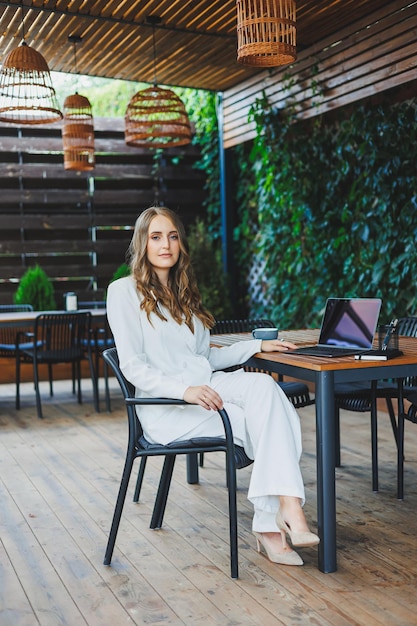 Een mooie vrouw in een wit klassiek pak werkt met een laptop op een open terras in een café Een vrouw in stijlvolle zakelijke kleding werkt op afstand op een laptop in een zomercafé