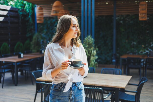 Een mooie vrouw in een wit overhemd en spijkerbroek drinkt koffie op het terras van een zomercafé en geniet van haar vakantie