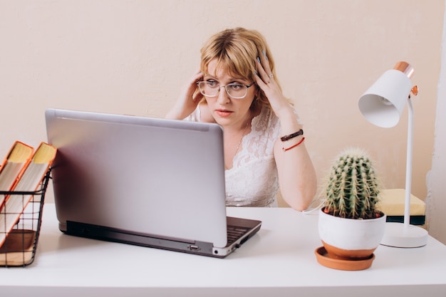 Een mooie vrouw in een witte blouse zit op kantoor en kijkt verbaasd naar de laptopmonitor