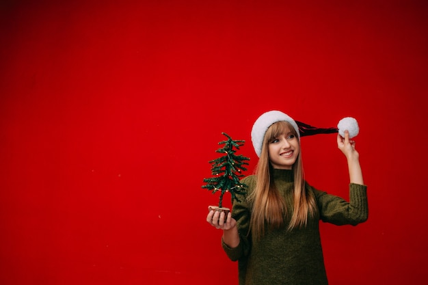 Een mooie vrouw met een kerstmuts houdt een kleine kerstboom in haar handen op een rode achtergrond