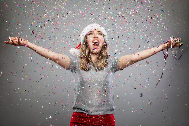 Foto een mooie vrouw met een kerstmuts in een regen van confetti lacht en geniet van de vakantie. traditionele winterevenementen., kerstmis en nieuwjaar. grijze achtergrond.