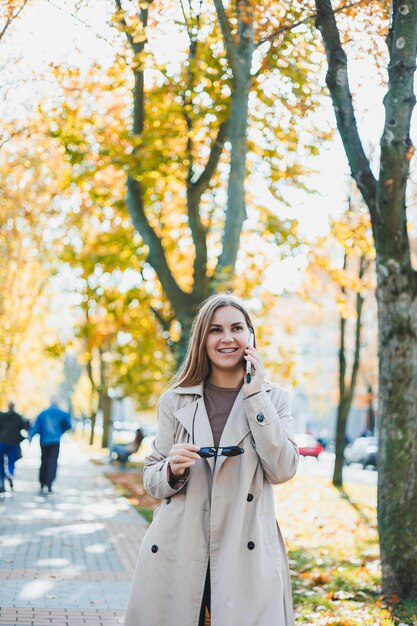 Een mooie vrouw met een zonnebril loopt in het herfstpark en praat aan de telefoon Vrolijk meisje op het herfststeegje in de stad