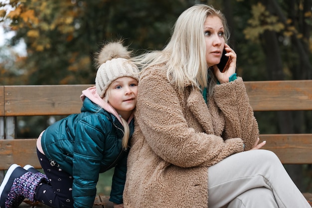 Een mooie vrouw praat aan de telefoon en haar dochtermeisje knuffelt haar in het herfstpark