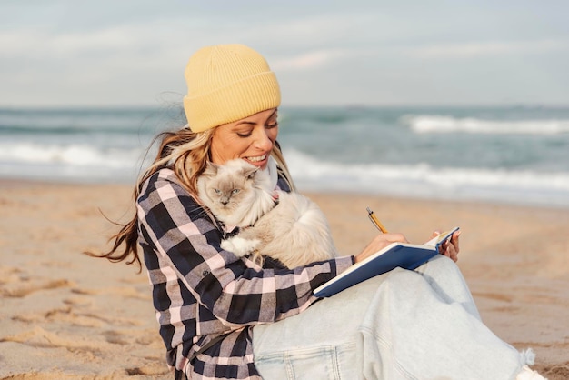 Een mooie vrouw schrijft in een dagboek en zit met haar kat op het zand